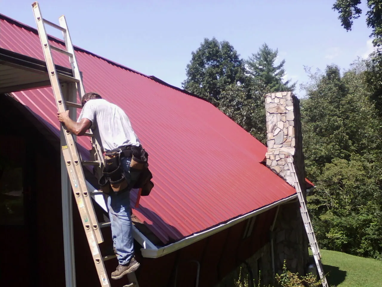 Roofer on a ladder beside a red metal roof and stone chimney — Soffit & Fascia Repair by Roofing Brothers in Ronda